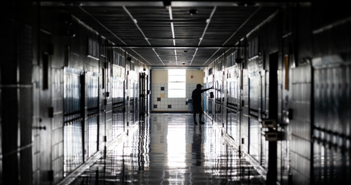 Dimly lit hallway of a high school, with lockers on either side, a man stands with his arms out touching a wall, conveying sadness.
