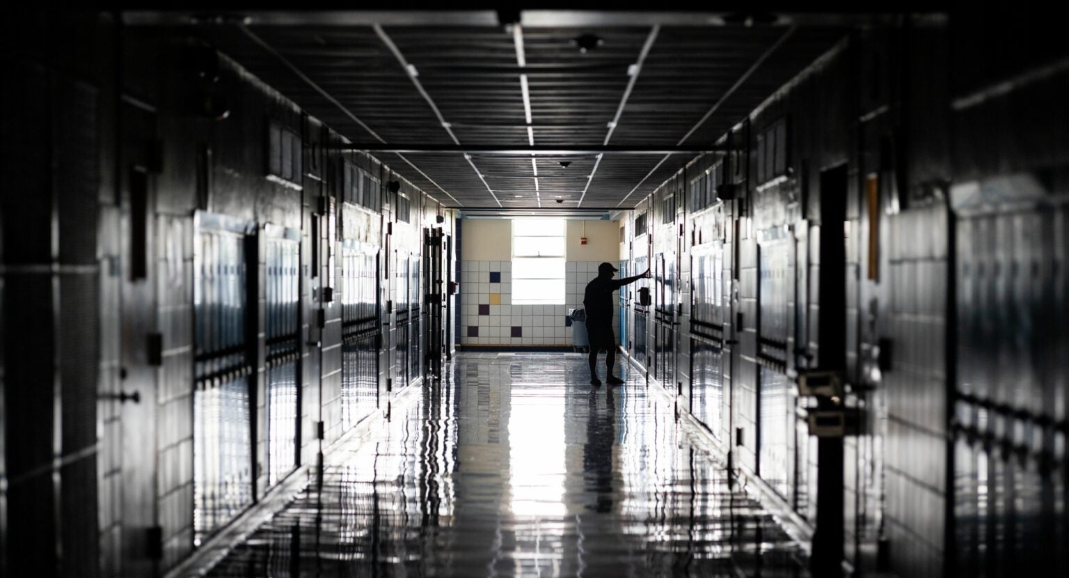 Dimly lit hallway of a high school, with lockers on either side, a man stands with his arms out touching a wall, conveying sadness.