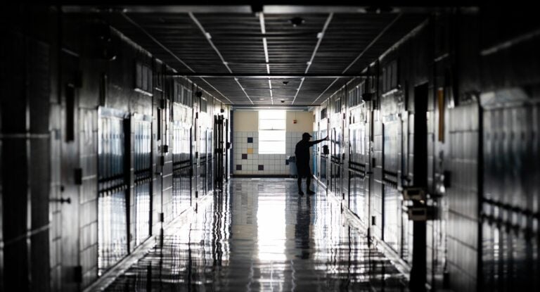 Dimly lit hallway of a high school, with lockers on either side, a man stands with his arms out touching a wall, conveying sadness.