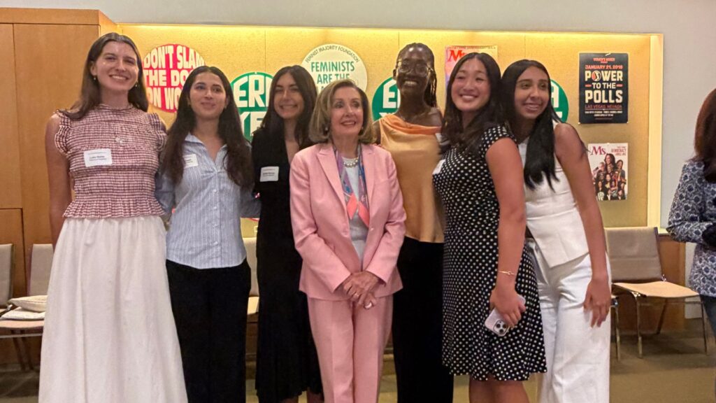 Luka Marks stands with Nancy Pelosi and group of women at Feminist Majority Foundation|Luka Marks Stands in front of wall holding up a poster that says Ms. This is what democracy looks like.