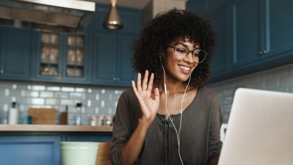 Woman raises hand to introduce herself on video call in front of her laptop sitting in her kitchen.