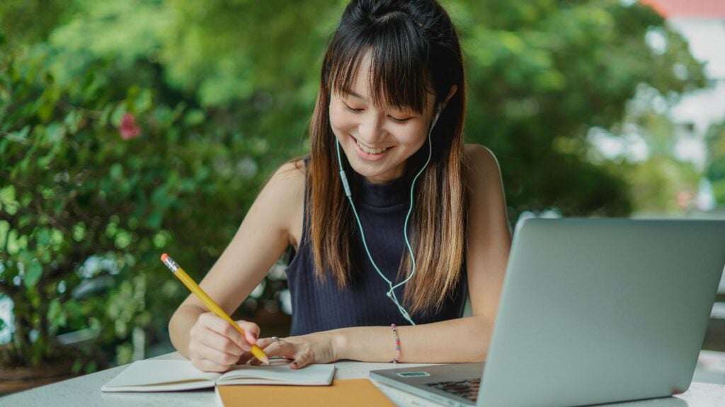 Female student sits at desk in front of a laptop and open notebook smiling as she writes her notes