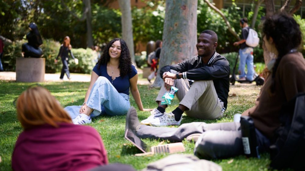4 students sit on the grass in the sculpture garden, Lydia Kelley sits and smiles at her friends in shot.|