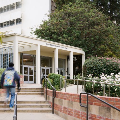 White building which is the east entrance of the UCLA Luskin school of public affairs building at UCLA with stairs leading up to the entrance, back of a student is walking up the stairs to the building entrance.