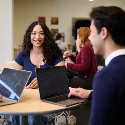 Luskin students sit around a table in the Luskin Commons smiling and talking with laptops on the table.