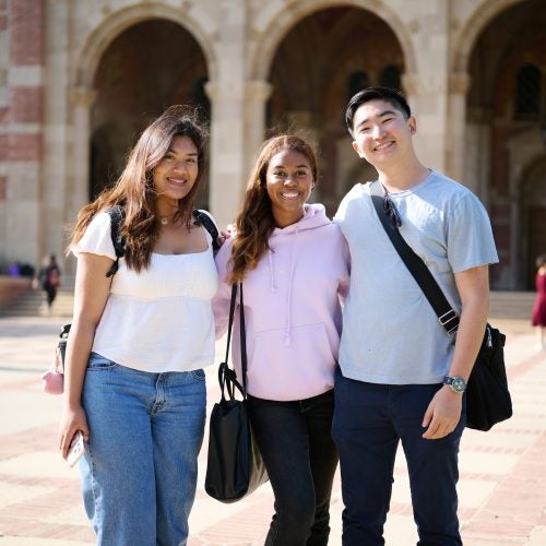 Three students stand shoulder to shoulder smiling at the camera in in front of Royce Hall on UCLA campus