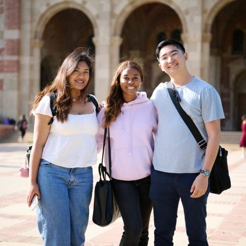 Three students stand shoulder to shoulder smiling at the camera in in front of Royce Hall on UCLA campus