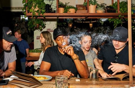 A man in a black T-shirt and baseball cap smokes a joint while sitting at the counter of a weed cafe in California.