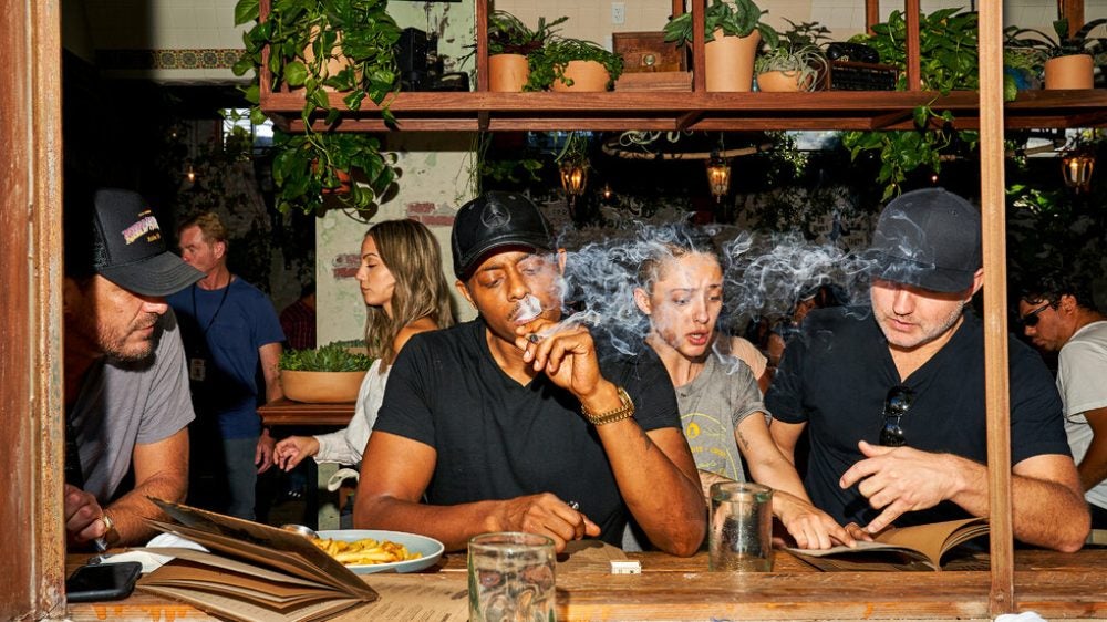 A man in a black T-shirt and baseball cap smokes a joint while sitting at the counter of a weed cafe in California.