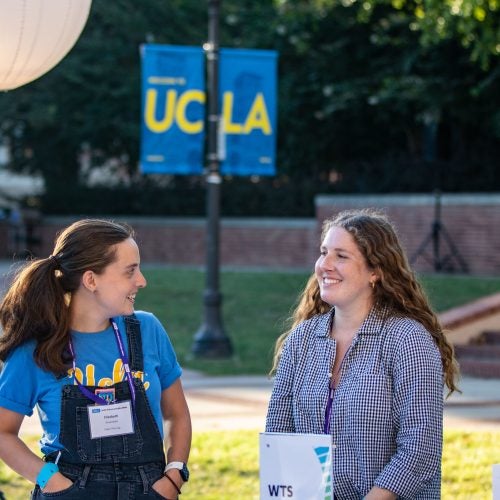 Two students at the annual Luskin Block Party standing in front of a UCLA sign, smiling smiling at each other on campus.