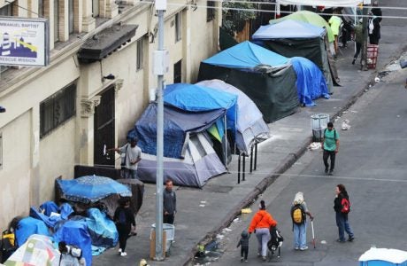 Blue tents set up on the sidewalk of a homeless encampment as some pedestrians walk by.