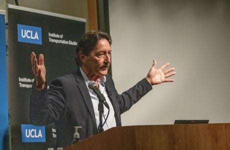 man at podium gestures with arms spread wide|man wearing glasses speaks at podium|woman with gray hair gestures|two men talk while seated in front of a projection screen