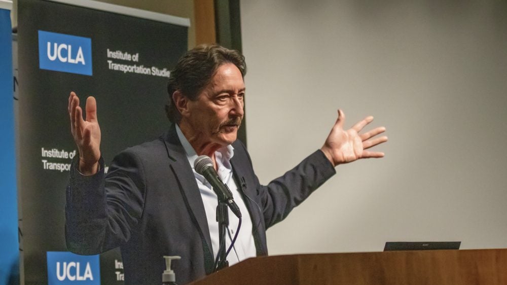 man at podium gestures with arms spread wide|man wearing glasses speaks at podium|woman with gray hair gestures|two men talk while seated in front of a projection screen