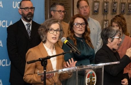 Woman in tan jacket at podium with others in background