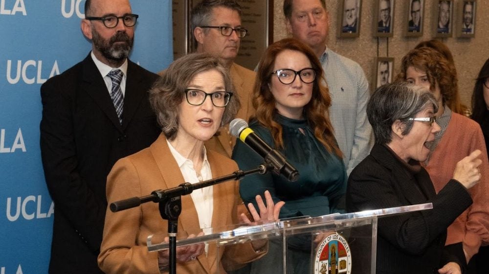 Woman in tan jacket at podium with others in background