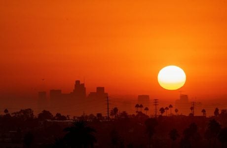 sun hangs low over city with sky looking orange Photo by johnemac72/iStock|woman in striped top speaks at podium while map is projected on a screen behind her|woman in dark shirt without sleeves speaks at a podium|man in blue shirt and glasses gestures at a podium while a woman listens int he background