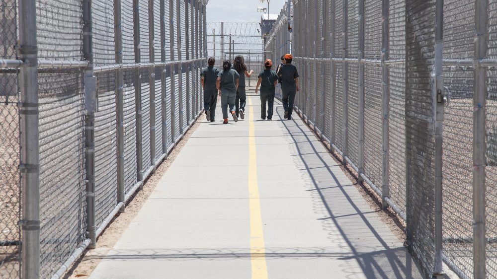 Group of people walking together in a fenced and gated ICE detention center.