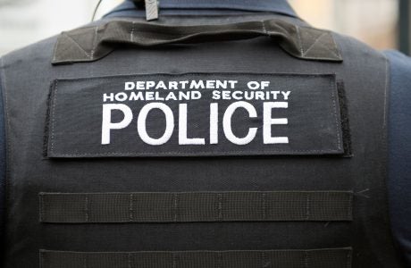 Photo of a shoulder and back of a person wearing a bullet proof vest with the words Department of Homeland Security Police written across the back in white letters.
