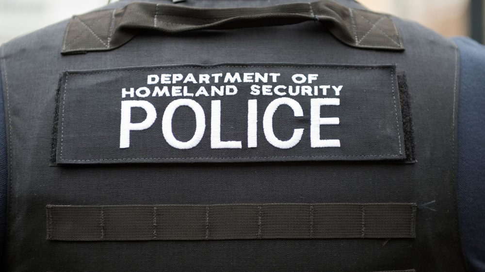 Photo of a shoulder and back of a person wearing a bullet proof vest with the words Department of Homeland Security Police written across the back in white letters.