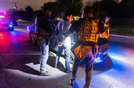 Immigration enforcement officers handcuff a man in the night, in front of a car in the middle of the road.