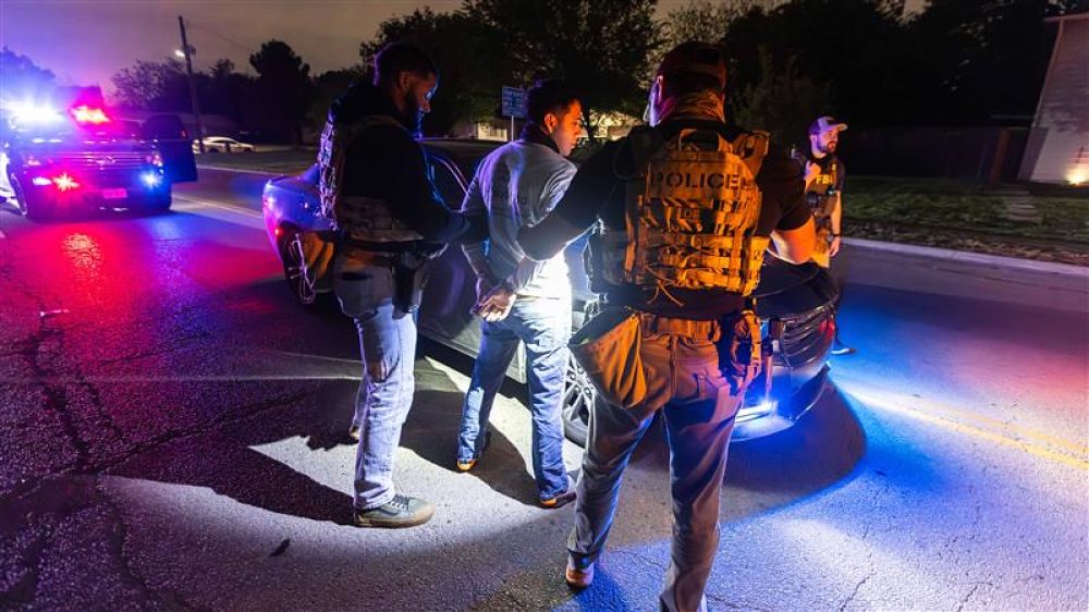 Immigration enforcement officers handcuff a man in the night, in front of a car in the middle of the road.