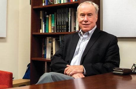 man sits with hands folded on lap and bookshelf in background
