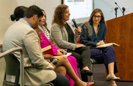 five people in business attire sit on a stage as a woman with curly brown hair speaks into a microphone