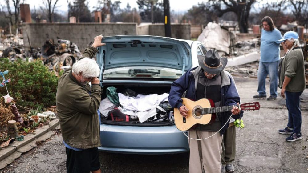 two people, one playing guitar, next to open car trunk amid fire zone