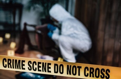 Crime scene yellow tape in the forefront with a detective in a white suit examining something on the ground in a forested area.