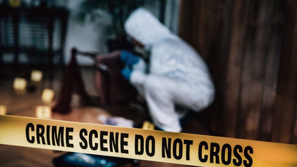 Crime scene yellow tape in the forefront with a detective in a white suit examining something on the ground in a forested area.