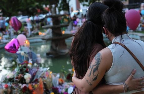 two women embracing in front of memorial of flowers and balloons