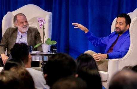 Two men in white chairs on stage in front of blue curtain