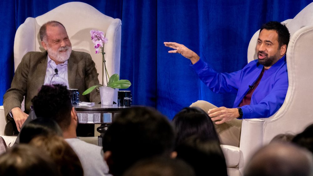Two men in white chairs on stage in front of blue curtain