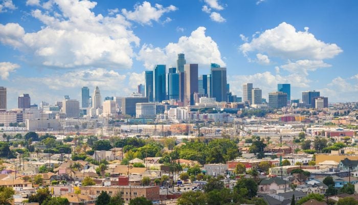 The Los Angeles skyline from a distance on a clear day: Tall buildings against a background of blue sky with clouds overhead. Smaller buildings and green trees stand in the foreground beneath the skyscrapers.