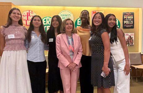 Luka Marks stands with Nancy Pelosi and group of women at Feminist Majority Foundation|Luka Marks Stands in front of wall holding up a poster that says Ms. This is what democracy looks like.