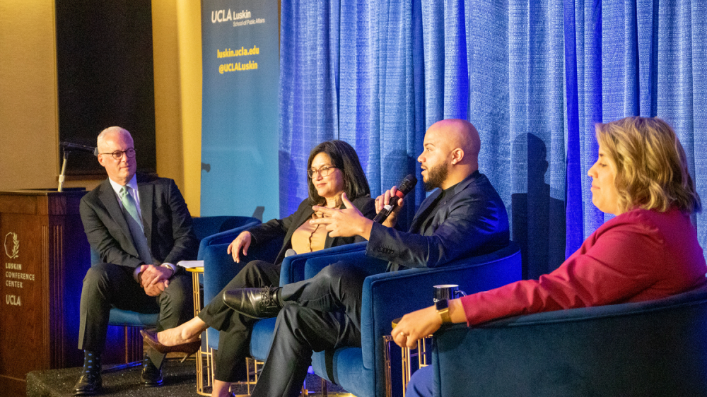 four people in business attire site on a stage and chat with a blue background behind them|||||
