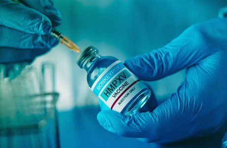 A healthcare or laboratory worker man, wearing blue surgical gloves, fills a syringe with a simulated monkeypox vaccine from a vial|man smiles as he stands beneath sign that designates office location for research hub