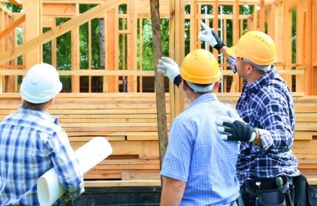 Three construction workers on a construction site with hard hats on pointing and talking together in front of a house with a wooden frame.|