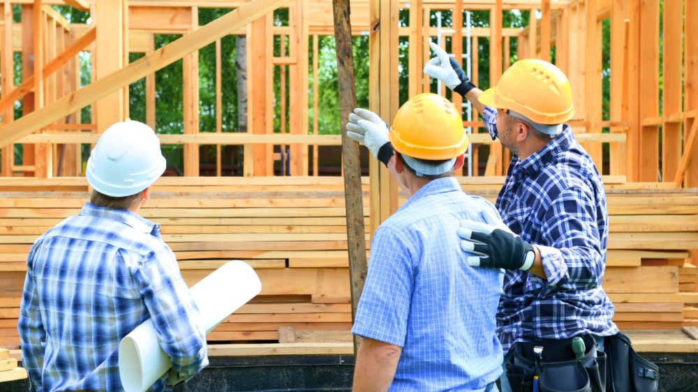 Three construction workers on a construction site with hard hats on pointing and talking together in front of a house with a wooden frame.|
