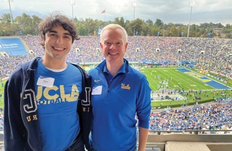 two young men smile in stands as UCLA football game goes on behind them