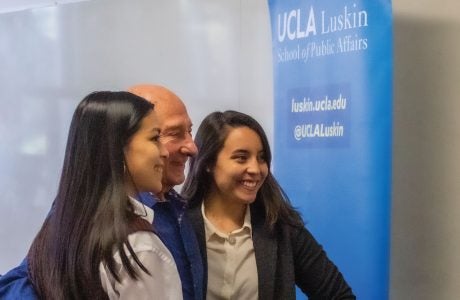 older man poses with two college students at UCLA event|people seated in foreground listen to speaker at podium while a screen shows an image of Martin Wachs|||