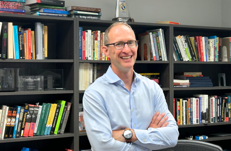 UCLA urban planning professor mike manville stands smiling in his office in front of a row of bookshelves.