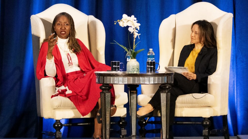 Two women are seated on stage, where a woman in red speaks and the other listens|woman in red shawl at podium gestures forward as she speaks|people seated at tables in a conference room watch a speaker at the front of the room speak at a podium|Two women are seated on stage, where a woman in red speaks and the other listens|woman in purple dress gestures at podium|Black woman leans forward as a woman holding a child speaks with her