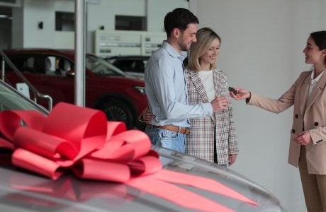 a new car at the dealership with a large red bow on the top, with three people in the background getting the keys to the brand new car.