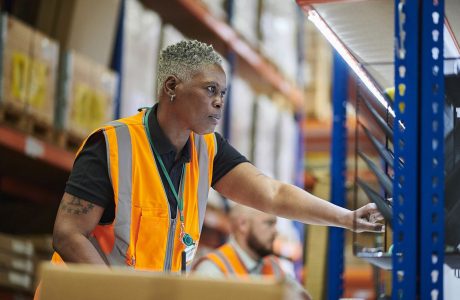 African American female warehouse worker wearing an orange vest doing her job.