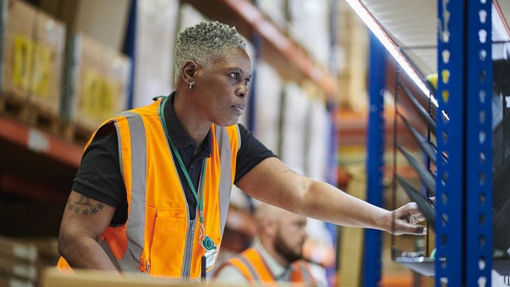 African American female warehouse worker wearing an orange vest doing her job.