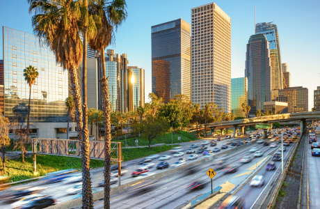 Blurred cars drive by on the freeway with palm trees in the foreground and tall buildings in the background on a sunny Los Angeles day