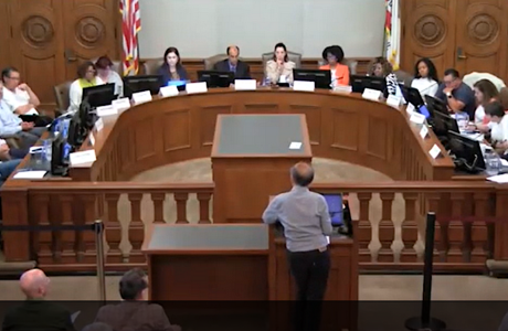 Man at podium addressing officials seated in semicircle