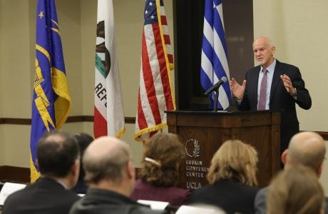 Former Greek prime minister George Papandreou speaking at UCLA. Greek flag and U.S. flag are behind him|woman with dark hair and glasses at podium