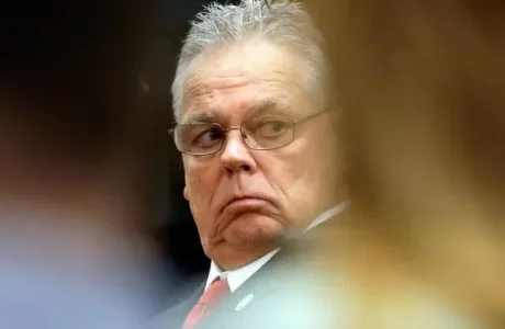 man in glasses and suit looks over his shoulder with a serious expression
